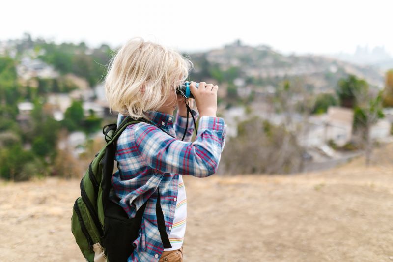 A young blonde boy stands outside in a park with hills and houses in the background. He is wearing a backpack and looking through binoculars.